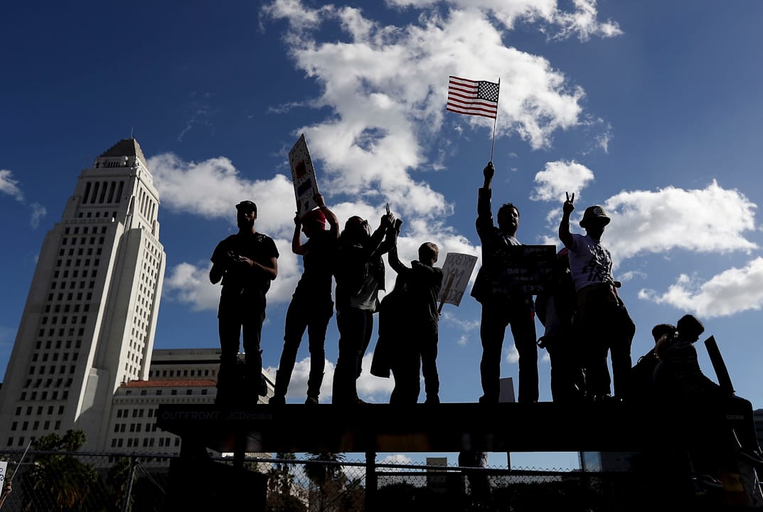 在美国加洲洛杉矶,有抗议者爬上巴士站上高举标语牌。