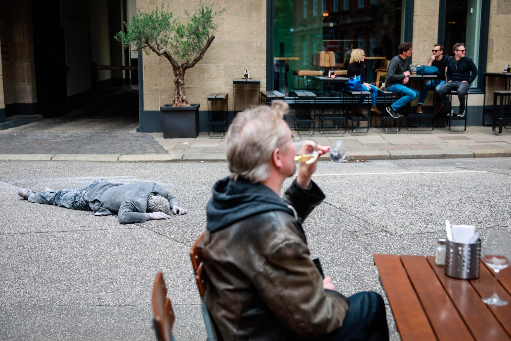 2017年7月5日,一名「1000 Figures」的參與者,在漢堡街頭的戶外酒吧前,躺在街道上,彷如死人。旁邊的酒客仍品嘗著杯中美酒。