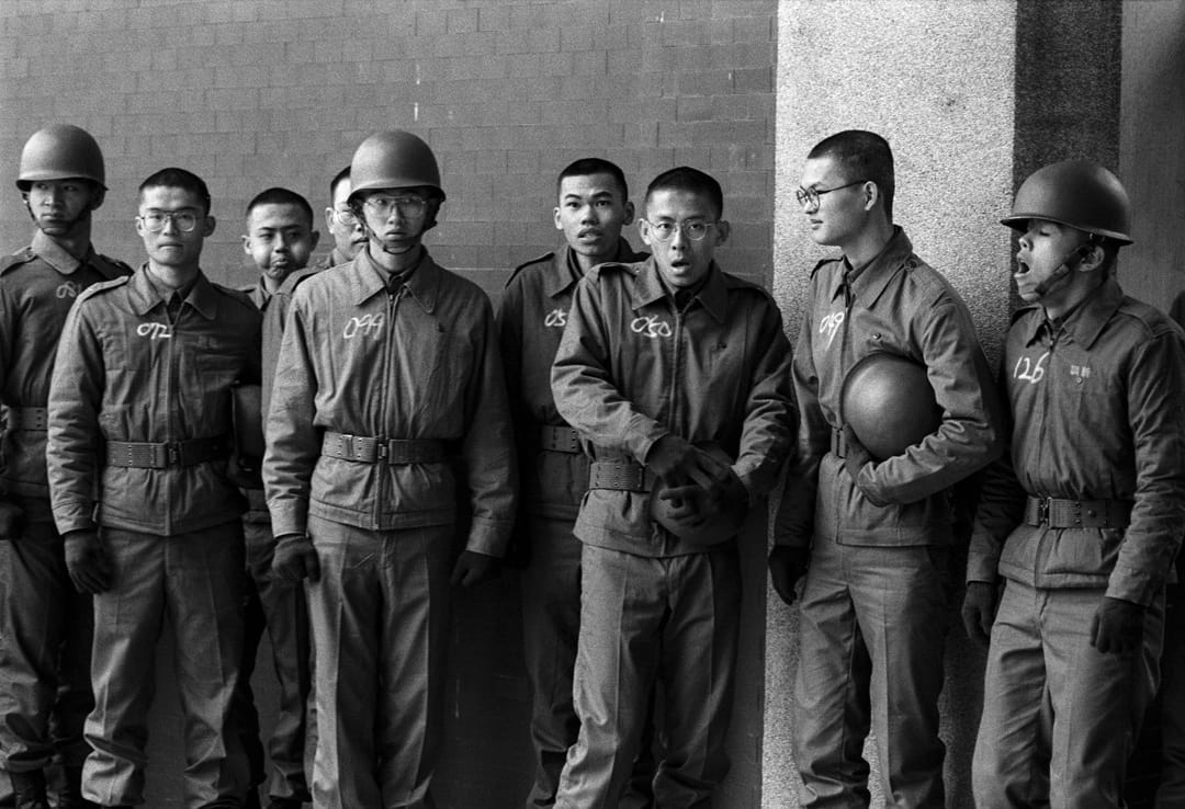 一群剛報到幾天，排隊等待拍攝大頭照的大專集訓學生，衣服上用粉筆寫着學號，個個表情是菜鳥，面對軍中環境的茫然。