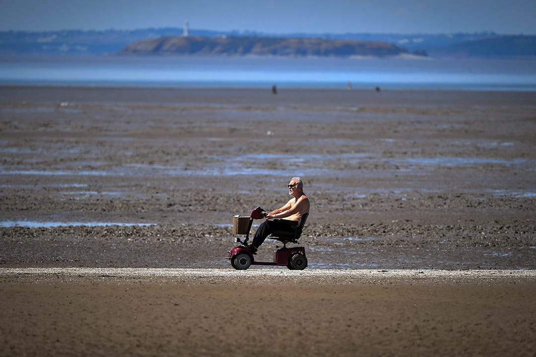 2018年7月15日,英國西部城鎮濱海韋斯頓,一名男子坐著電動代步車經過沙灘。