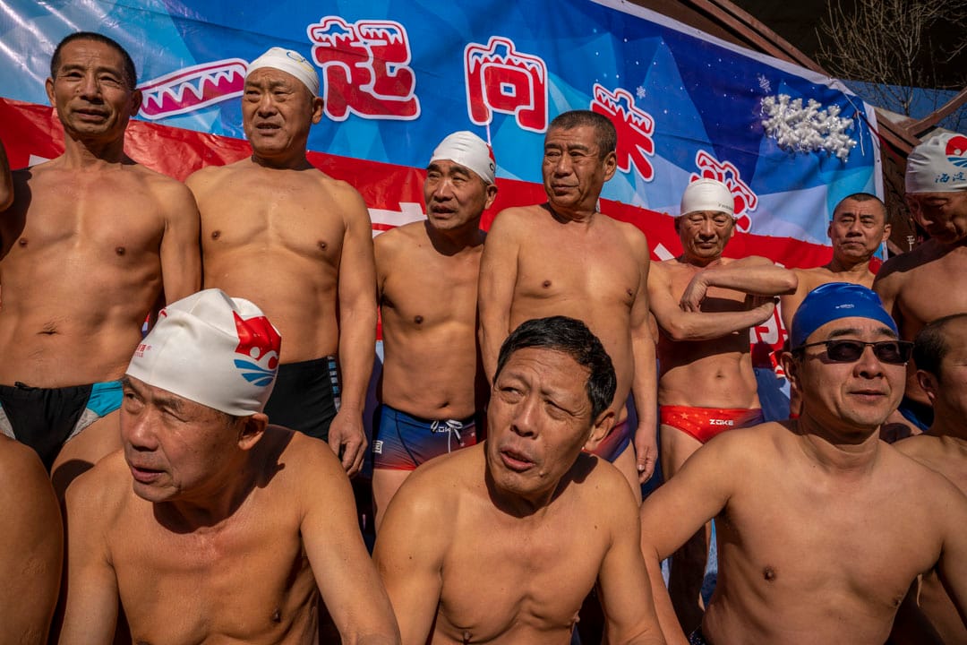 2022年2月3日中国北京，一群老人在北京冬奥会横幅前拍照，然后冰冻的河中游泳。