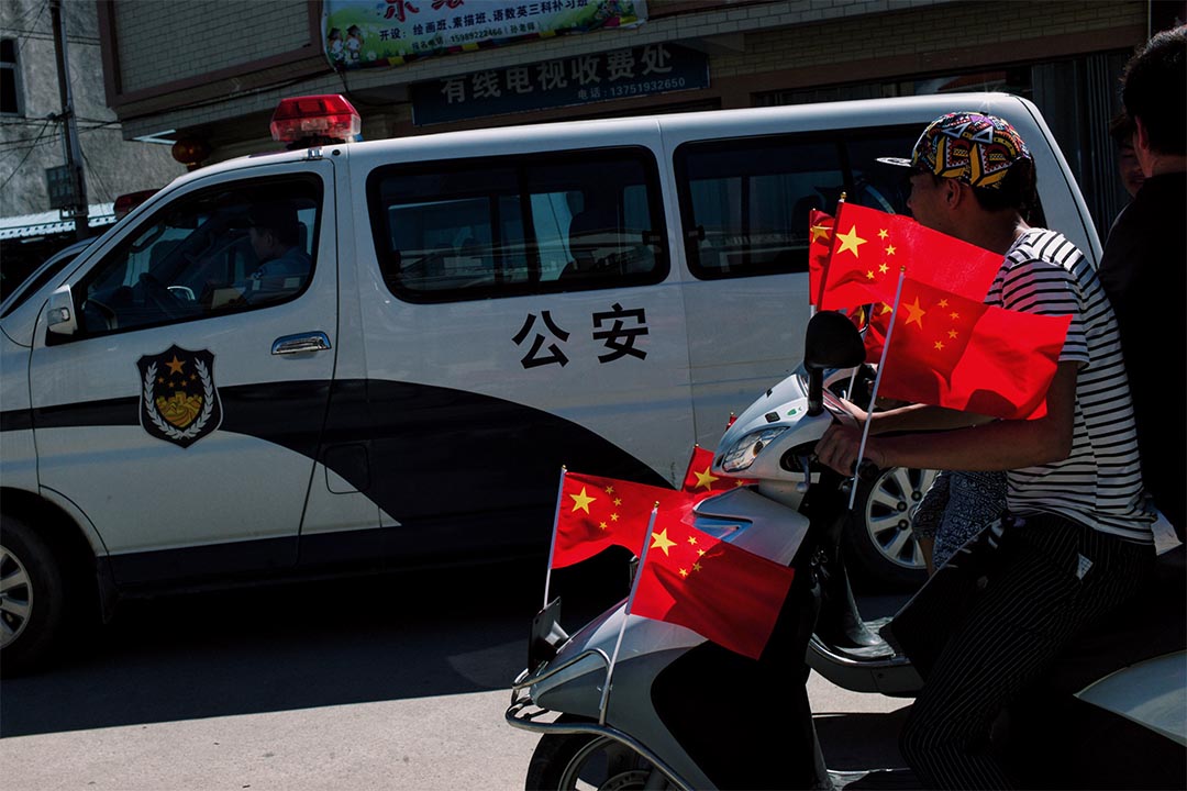 参与游行的村民乘着插满国旗的摩托车，驶经停泊于村口的公安车辆。