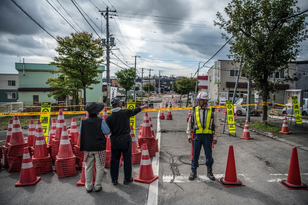 2018年9月8日，日本札幌清田区，道路在地震中严重损毁，需封闭进行维修，有道路工人在封锁线引导市民到安全地方，两名途人远观室内的损毁情况。