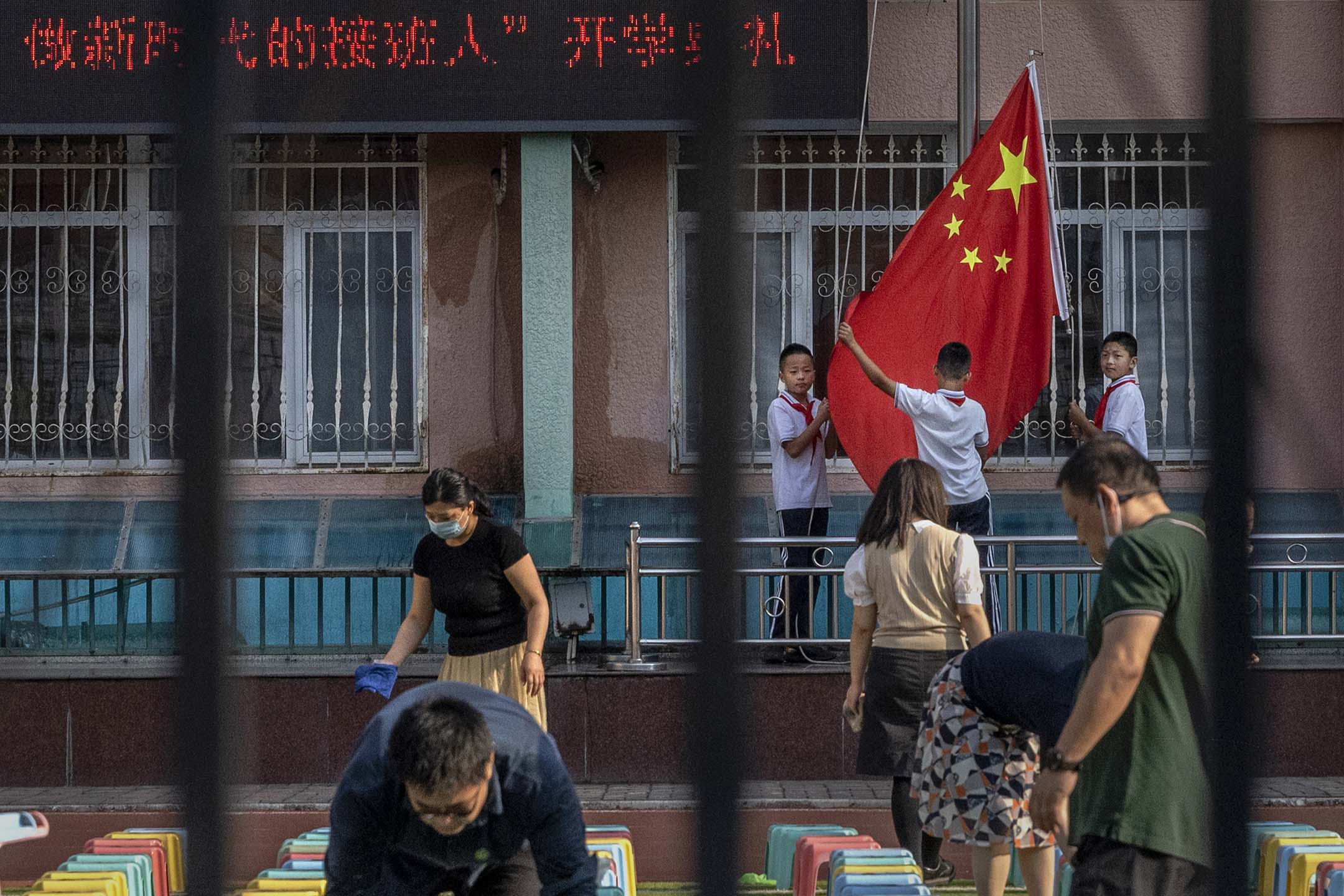 2021年9月1日中国北京，一所小学新学年第一天的开学仪式前，学生们准备一面国旗，老师们正安排凳子。