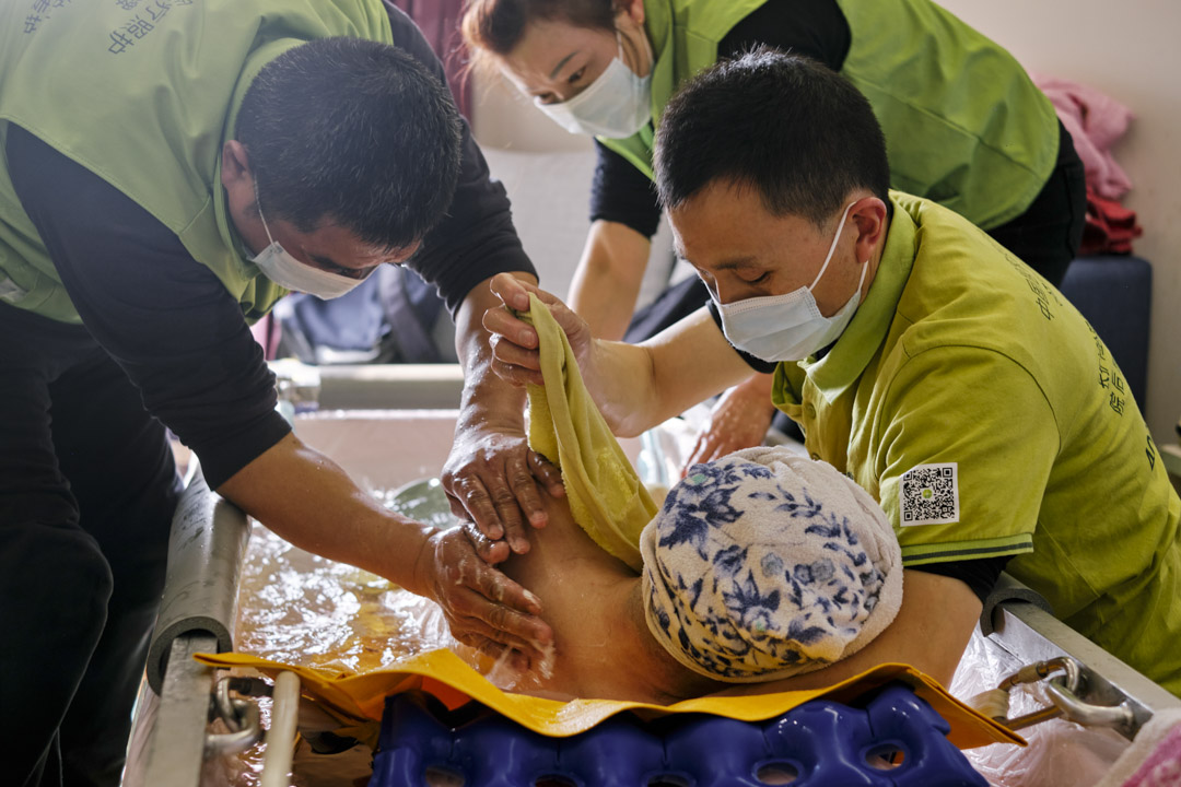 老人翻身洗背时需要两个助浴师一起，陈菊清侧卧在助浴师手臂上。