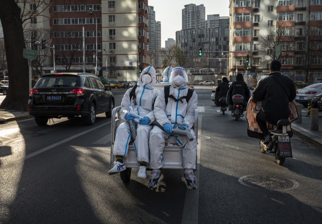 2022年12月9日，中國北京，兩名穿保護衣的防疫人員坐在三輪車的車尾上前往為疫區消毒。